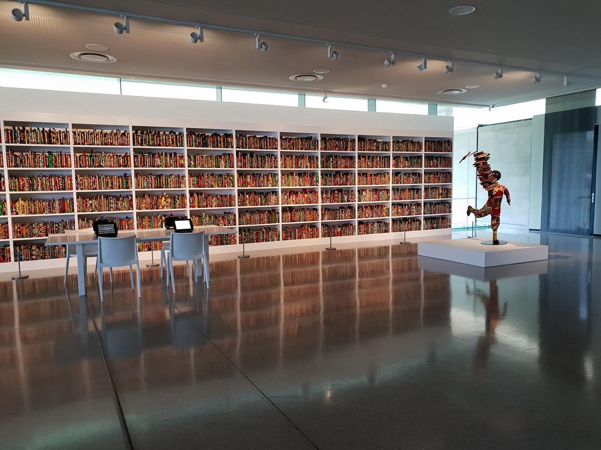 First-floor gallery with a full wall of shelved archive volumes and a single sculpture on a plinth