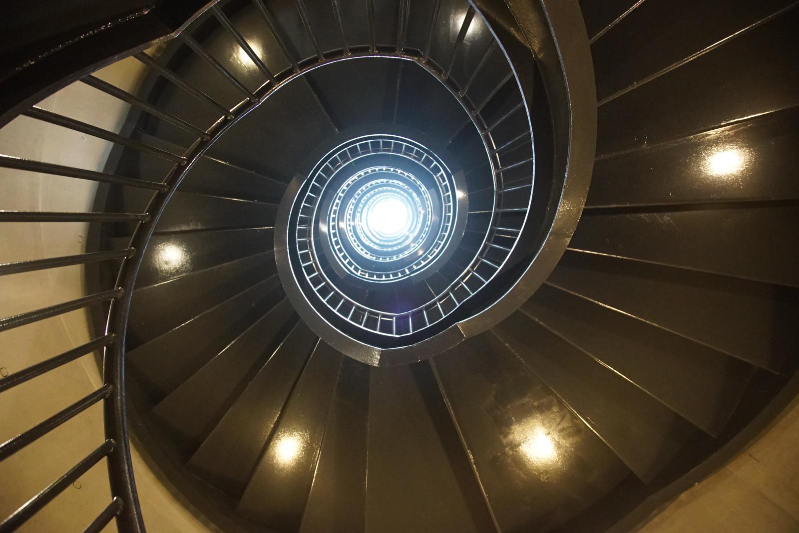 Spiral staircase inside a silo cylinder rising toward a circular skylight